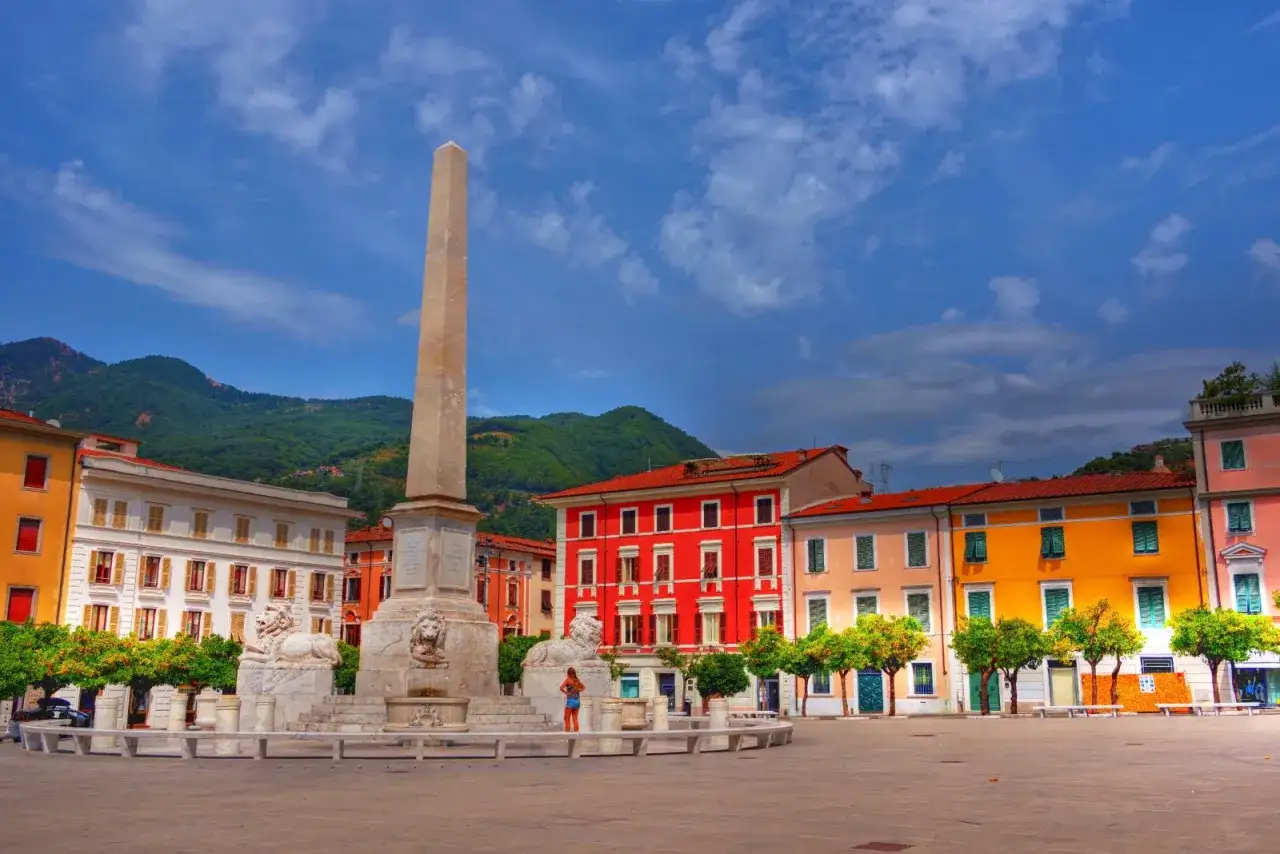 Piazza con obelisco e leoni, case colorate e montagne sullo sfondo. Un'idea di cosa vedere a Marina di Massa.