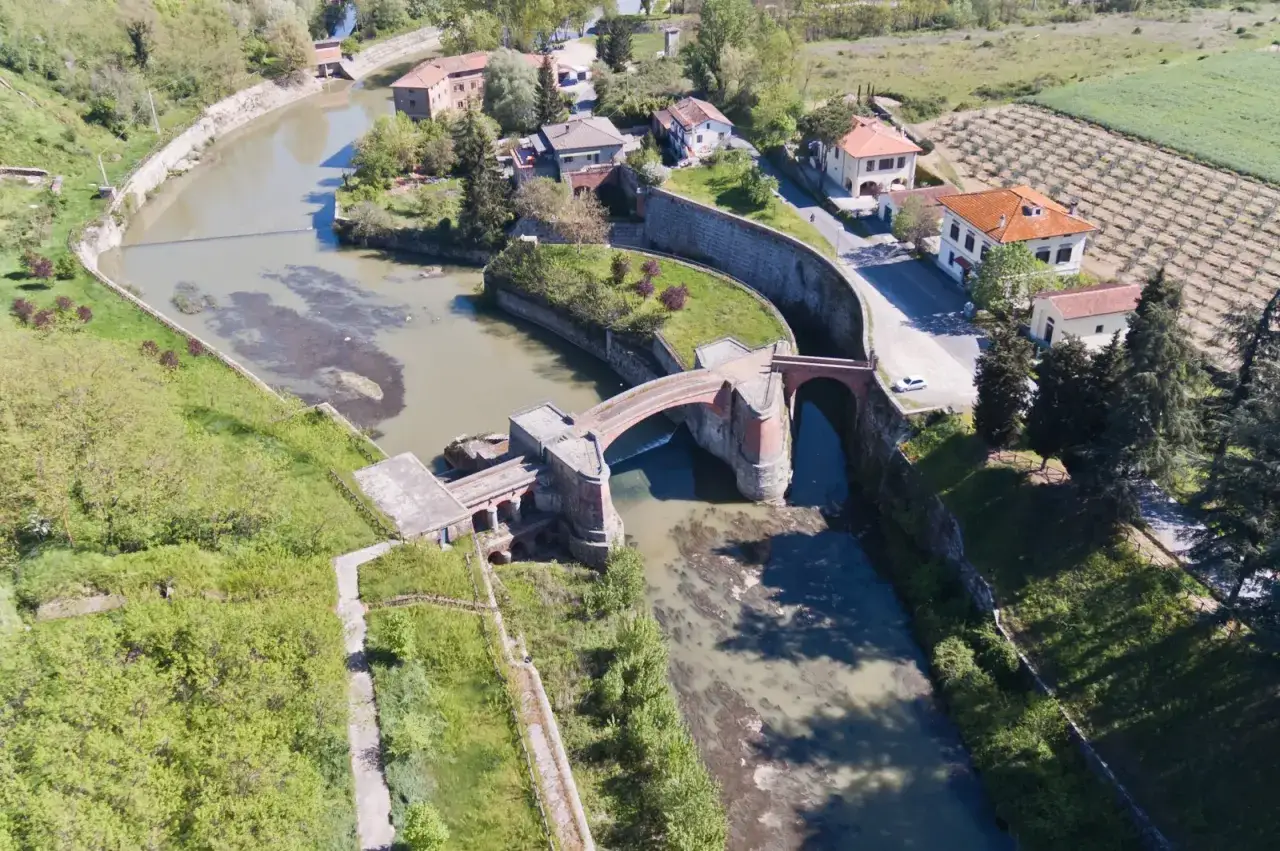 Ponte in mattoni sul fiume, con case e vigneti. Il sentiero della bonifica serpeggia tra il verde.