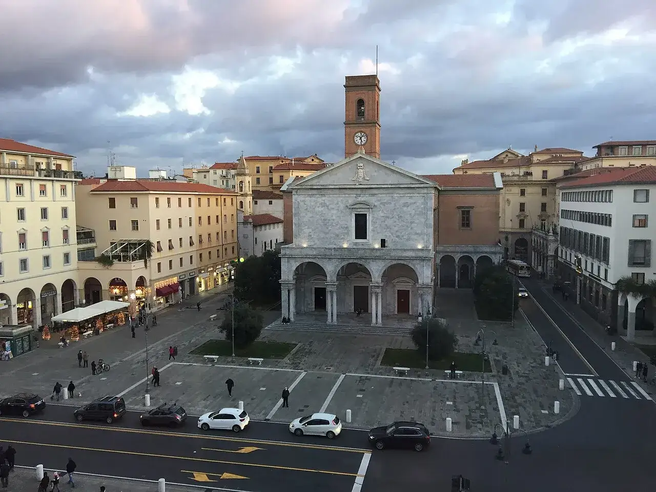 Il duomo di Livorno, con la sua facciata in marmo e il campanile con orologio, domina la piazza. Edifici color pastello e traffico cittadino completano la scena.