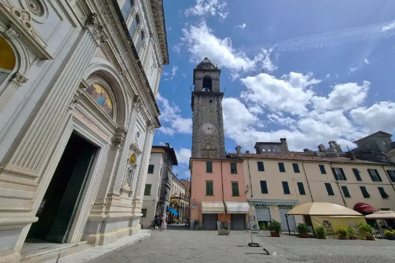 Piazza a Casola in Lunigiana con la chiesa imponente, la torre dell'orologio e case colorate sotto un cielo azzurro con nuvole bianche.