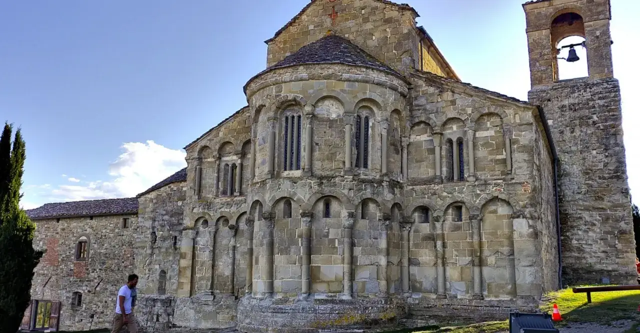 La Pieve di Romena, un gioiello romanico in pietra, si erge maestosa sotto un cielo azzurro. Un uomo passeggia, ammirando la sua antica bellezza.