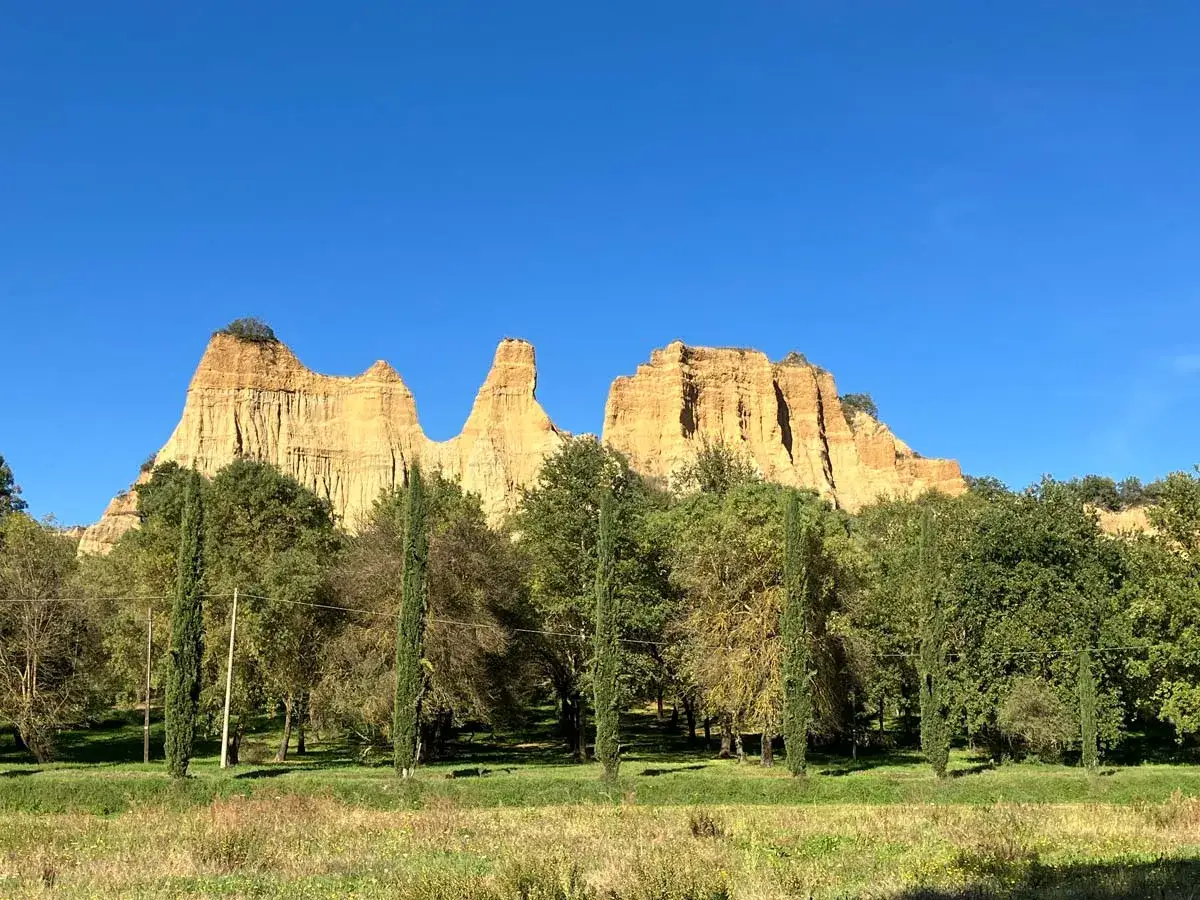 Paesaggio con calanchi dorati che ricordano il sentiero dell'acqua zolfina, circondati da alberi verdi e un cielo azzurro intenso.