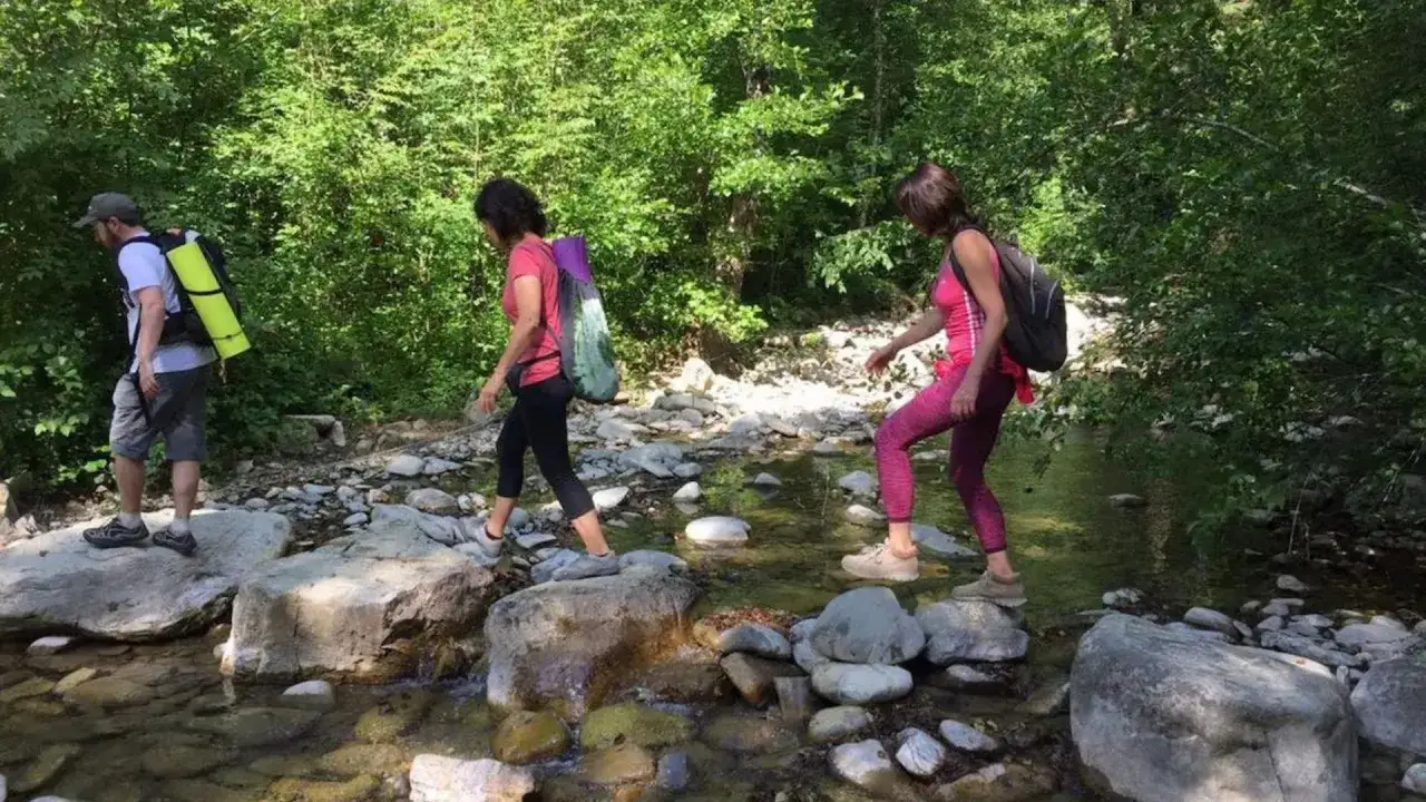 Tre escursionisti attraversano un torrente su pietre, immersi nel verde dei monti rognosi.