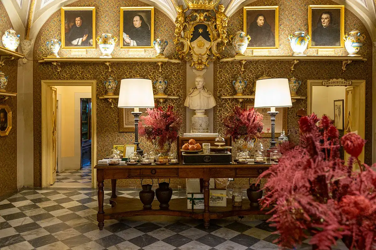 Interno elegante con ritratti antichi, vasi decorati e un tavolo con prodotti. L'atmosfera evoca la pi&ugrave; antica farmacia di Firenze.