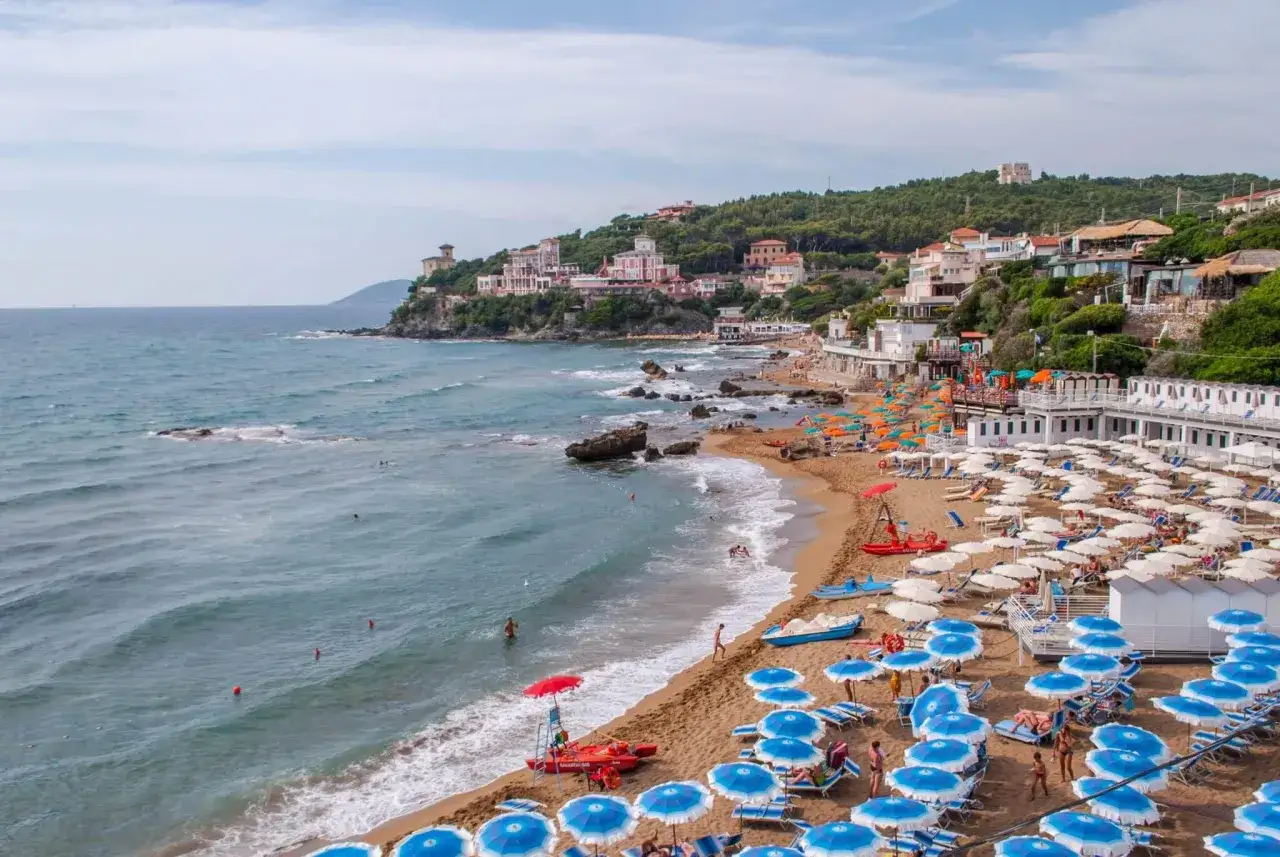 Spiaggia affollata a Castiglioncello, con ombrelloni blu e bianchi, lettini, stabilimenti balneari e ville sulla collina. Cosa vedere: il mare, la costa e il borgo.