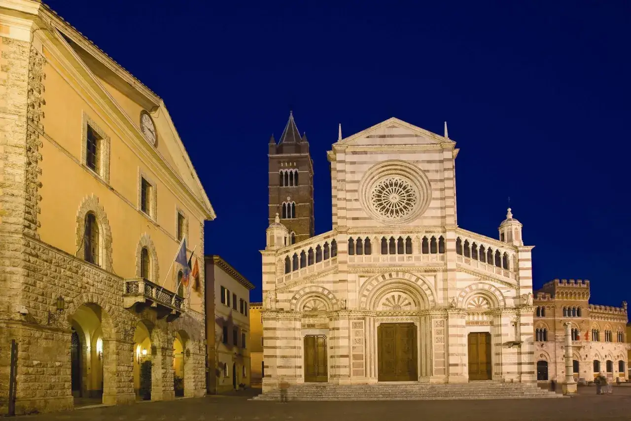 Il Duomo di Grosseto illuminato di notte, con la sua facciata a strisce bianche e rosa e il rosone circolare. Accanto, un edificio giallo con un orologio.