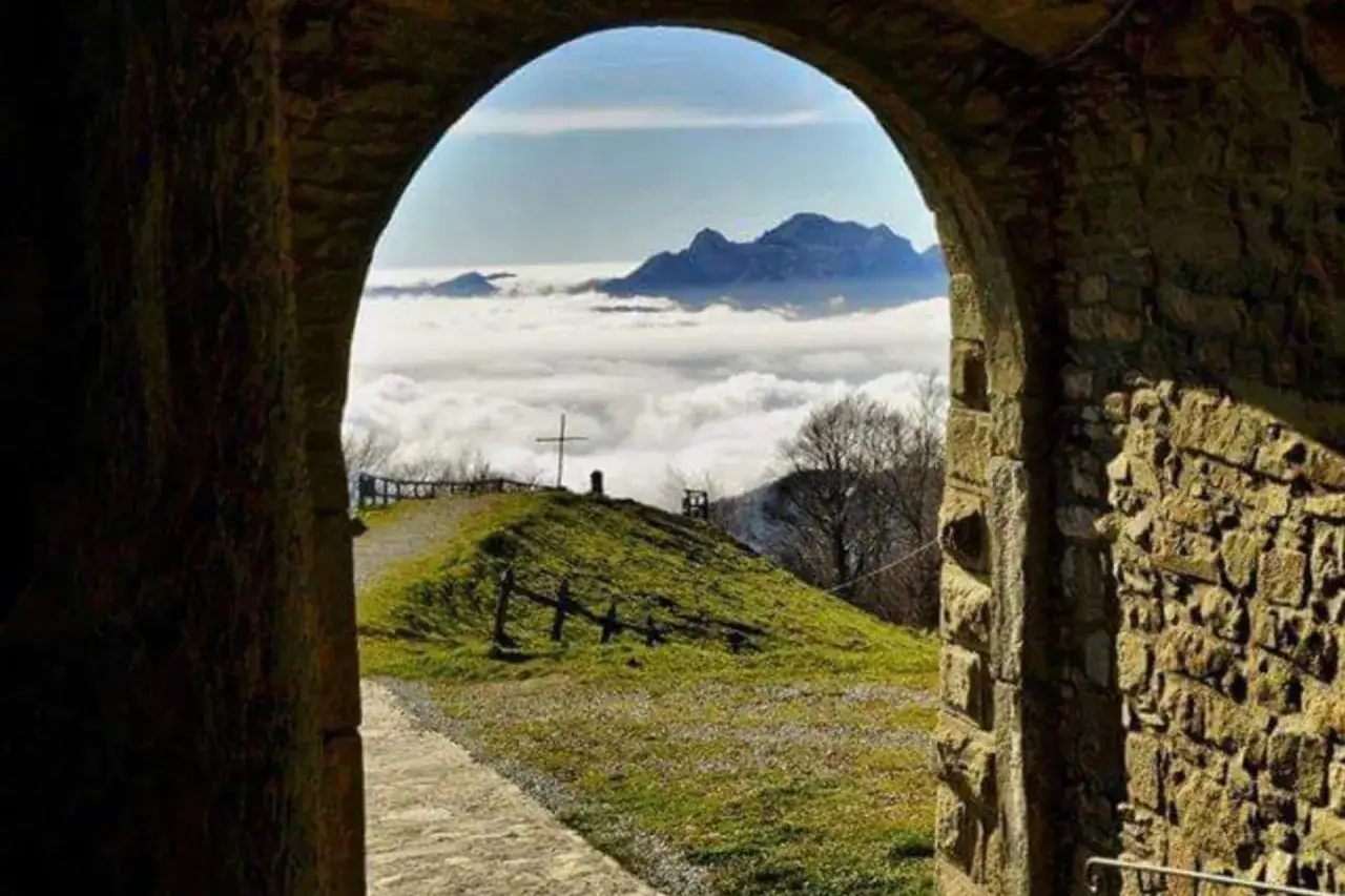 Un arco in pietra incornicia un paesaggio montano sopra un mare di nuvole. Una croce si erge su una collina verde, un punto di riferimento lungo la via Matildica del Volto Santo.