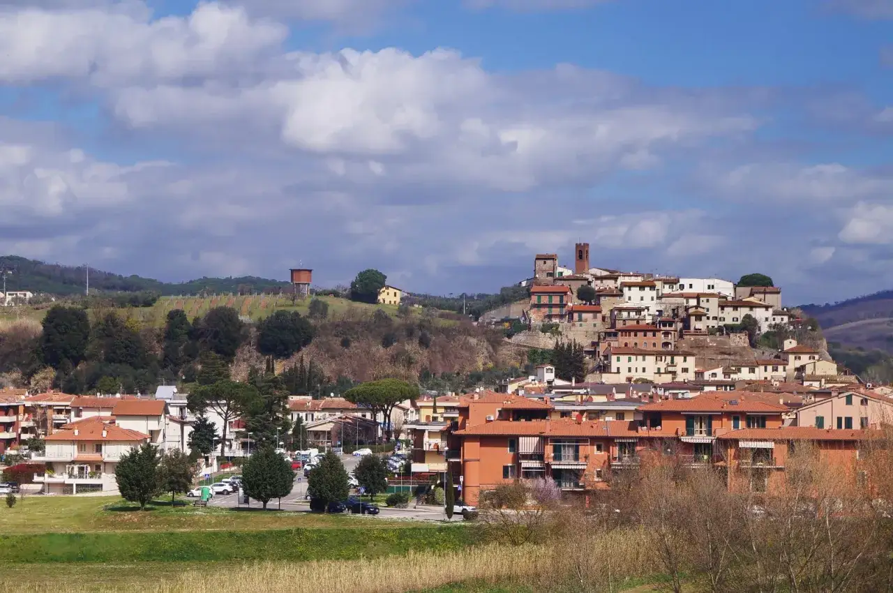 Panoramica di un borgo collinare toscano, con case dai tetti rossi e una torre medievale. Si intravede il limite del paese tra la campagna e le abitazioni.