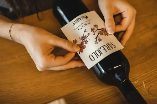Mani che applicano un'etichetta su una bottiglia di vino. L'etichetta indica "Toscana", la regione di origine del vino Sangiovese.