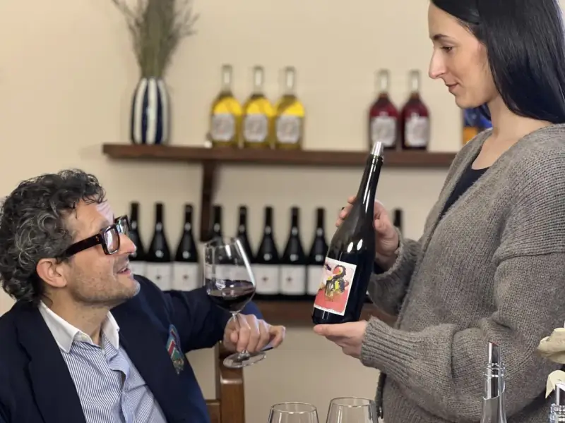 Uomo con occhiali e bicchiere di vino rosso, donna presenta bottiglia. Degustazione vini Arezzo in cantina.