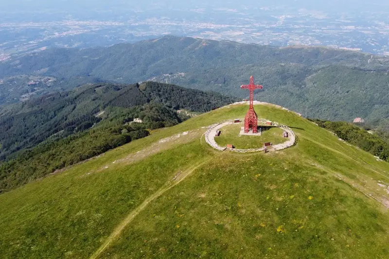 La croce del Pratomagno domina un paesaggio verdeggiante, con montagne boscose e una città in lontananza.