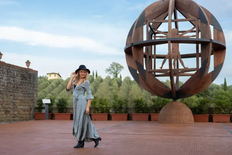 Donna con cappello nero ammira l'Uomo Vitruviano in una sfera di legno, un'opera iconica da vedere a Vinci, con colline verdi sullo sfondo.