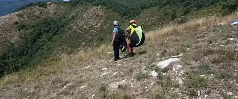 Due parapendisti si preparano al decollo sulla **Valle del Serchio**, con un paesaggio verdeggiante alle spalle.