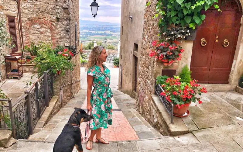 Una donna con un cane passeggia in un vicolo fiorito di Civitella Val di Chiana, con una vista panoramica sullo sfondo.
