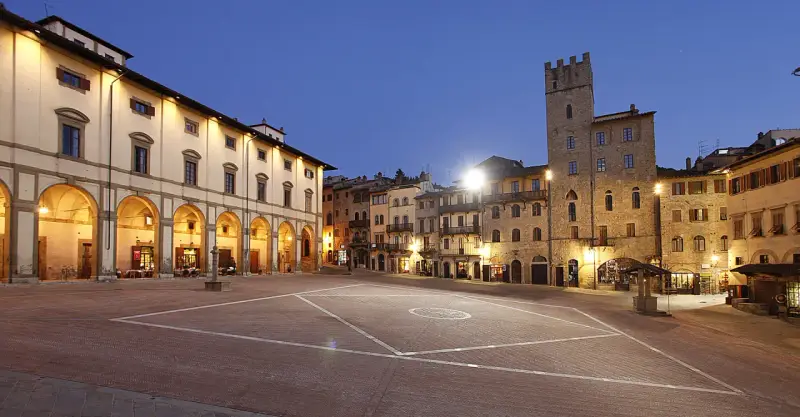 Piazza illuminata di Arezzo al crepuscolo, con portici, negozi e una torre medievale. Qui la vita è bella.