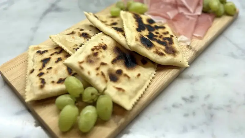Tortelli alla lastra casentino, dorati e leggermente bruciacchiati, serviti con uva fresca e affettati di prosciutto su un tagliere di legno.
